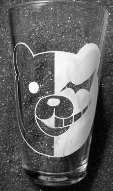 An etched pint glass with Monokuma's face on it. The left half is white with a batwing eye and smiling teeth. The right side is clear and has a round eye and gentle smile. 