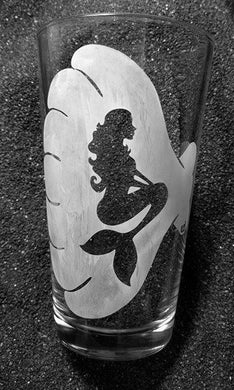 A pint glass with an etched mermaid design. The silhouette of a mermaid with long curly hair is sitting, framed by a large scalloped shell. 