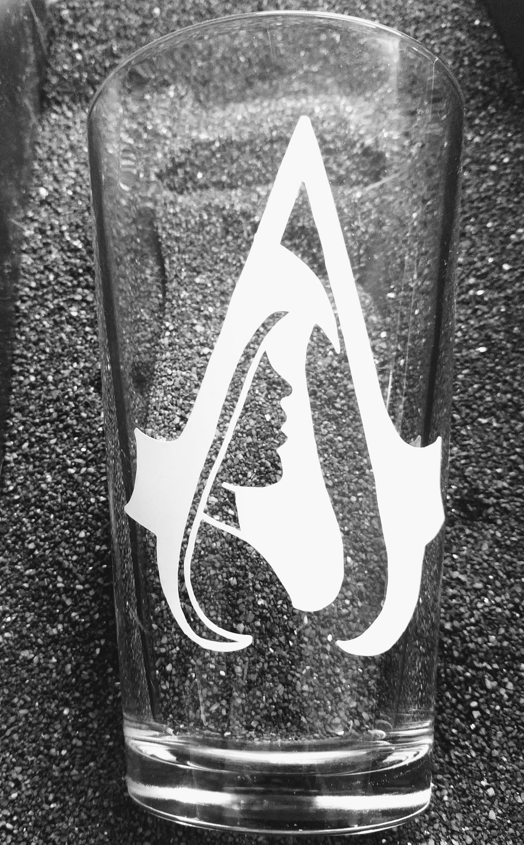 A pint glass with the Assassin's Creed A on it. Inside of the A is the profile of a man wearing an Assassins Creed hood. 