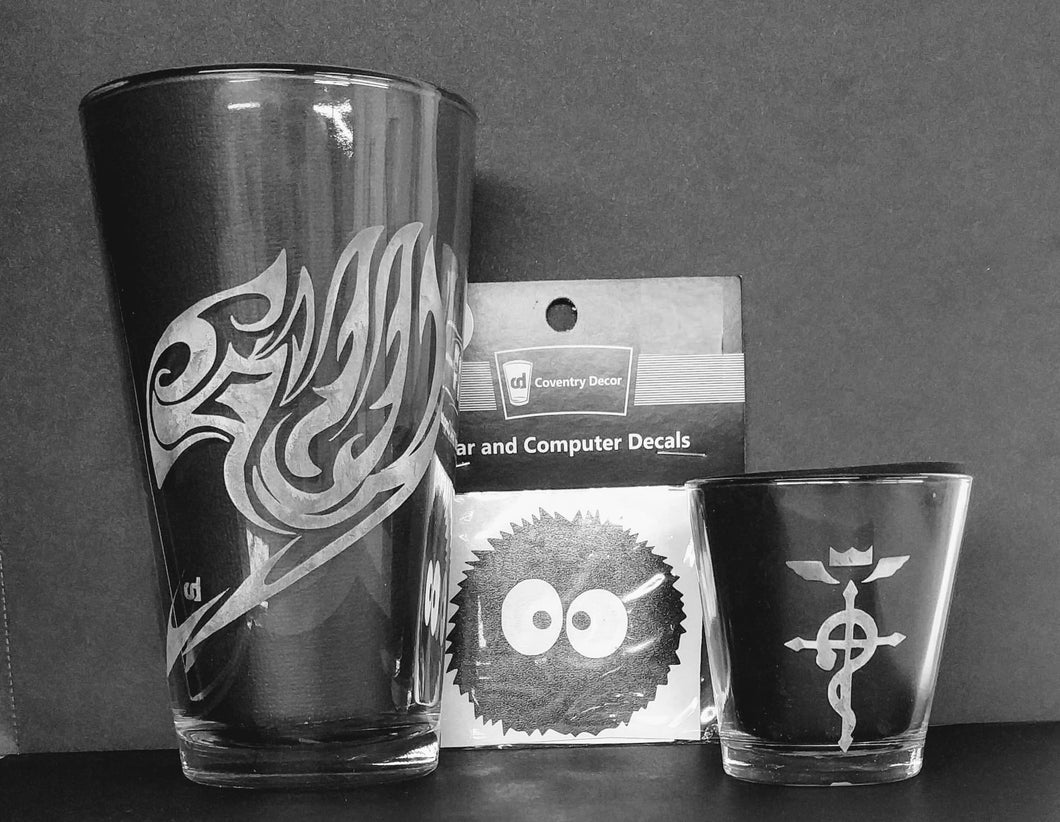 A Fairy Tail pint glass, a Soot Sprite decal, and a Fullmetal Alchemist shot glass against a black background. 