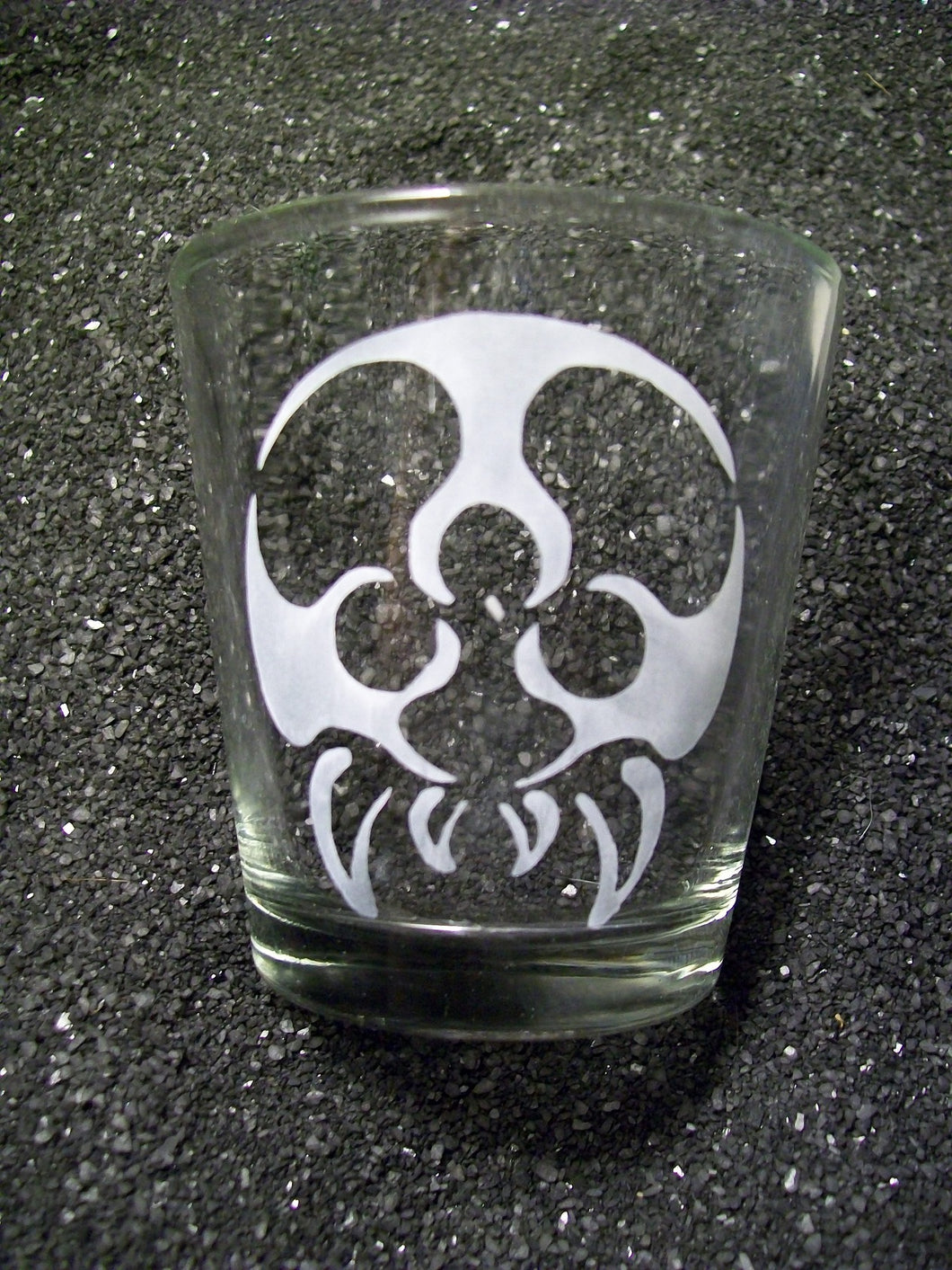 An extra large shot glass that is etched with a tattoo-style metroid monster. It has a round body (no head) with three eyeballs and four fangs. 