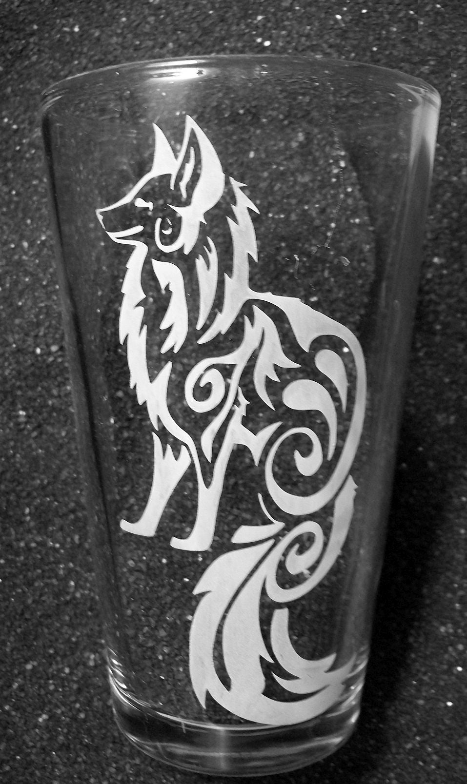 A pint glass with a tattoo-style fox design. The fox is seen from the side, sitting with its tail hanging down. Swirls decorate the fox's body. 