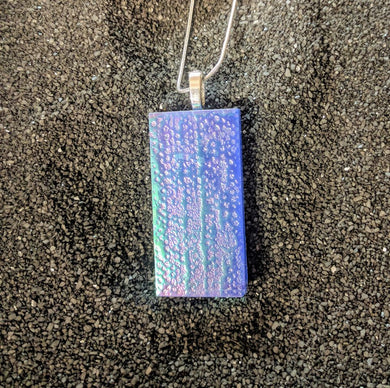 A rectangular glass pendant hangs from a silver chain. The pendant is blue with a slightly bumpy texture and a green holographic sheen. 