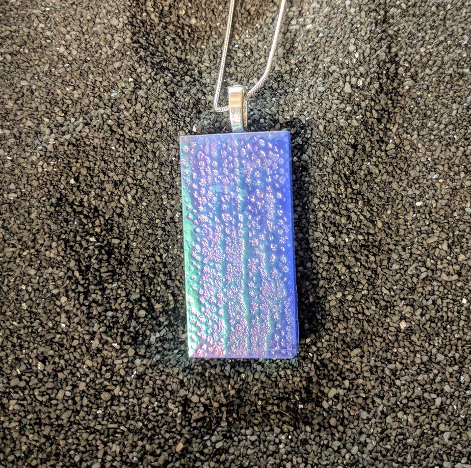 A rectangular glass pendant hangs from a silver chain. The pendant is blue with a slightly bumpy texture and a green holographic sheen. 